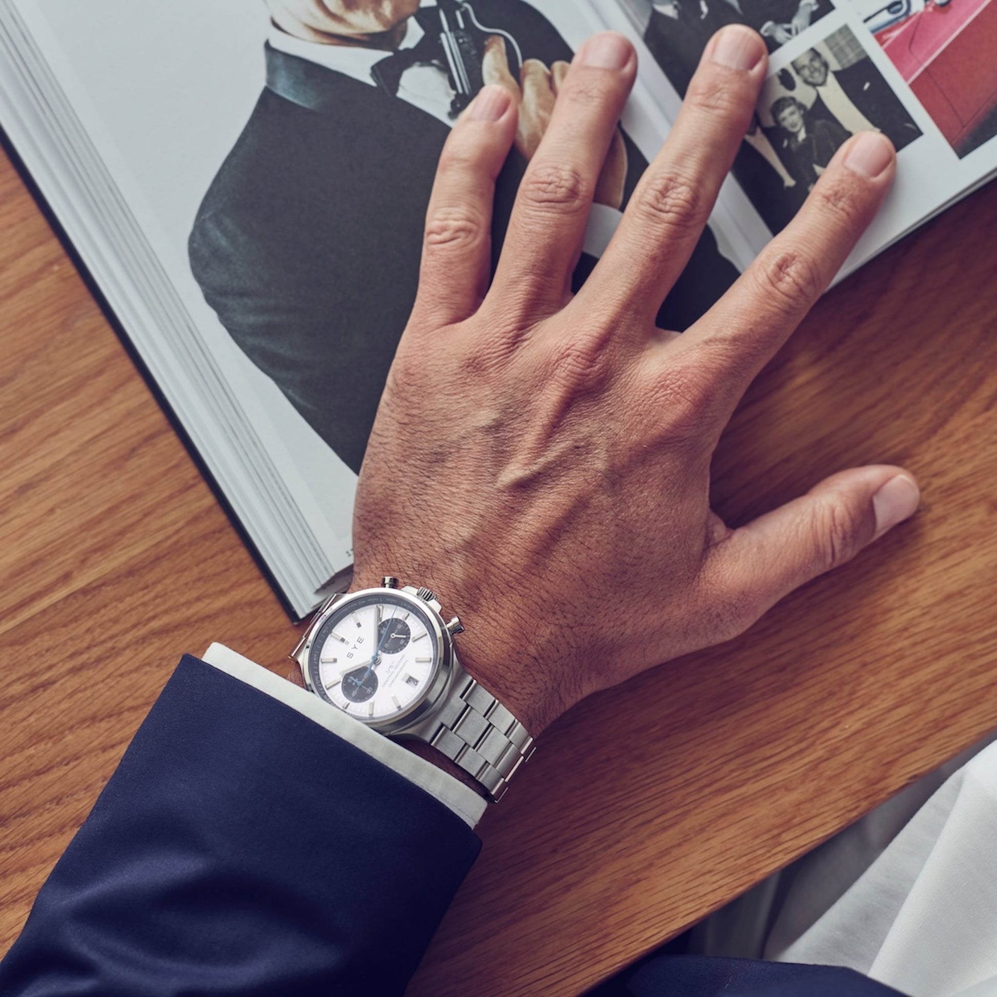 Hand wearing a silver watch on a wooden surface with a magazine in the background