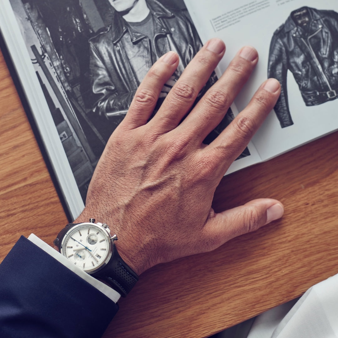 Hand wearing a watch on a wooden surface with a magazine open to a leather jacket page.