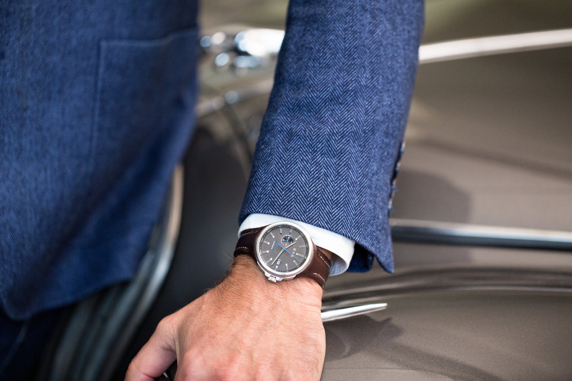 Hand wearing a watch with a blue suit sleeve, sitting on a car seat.