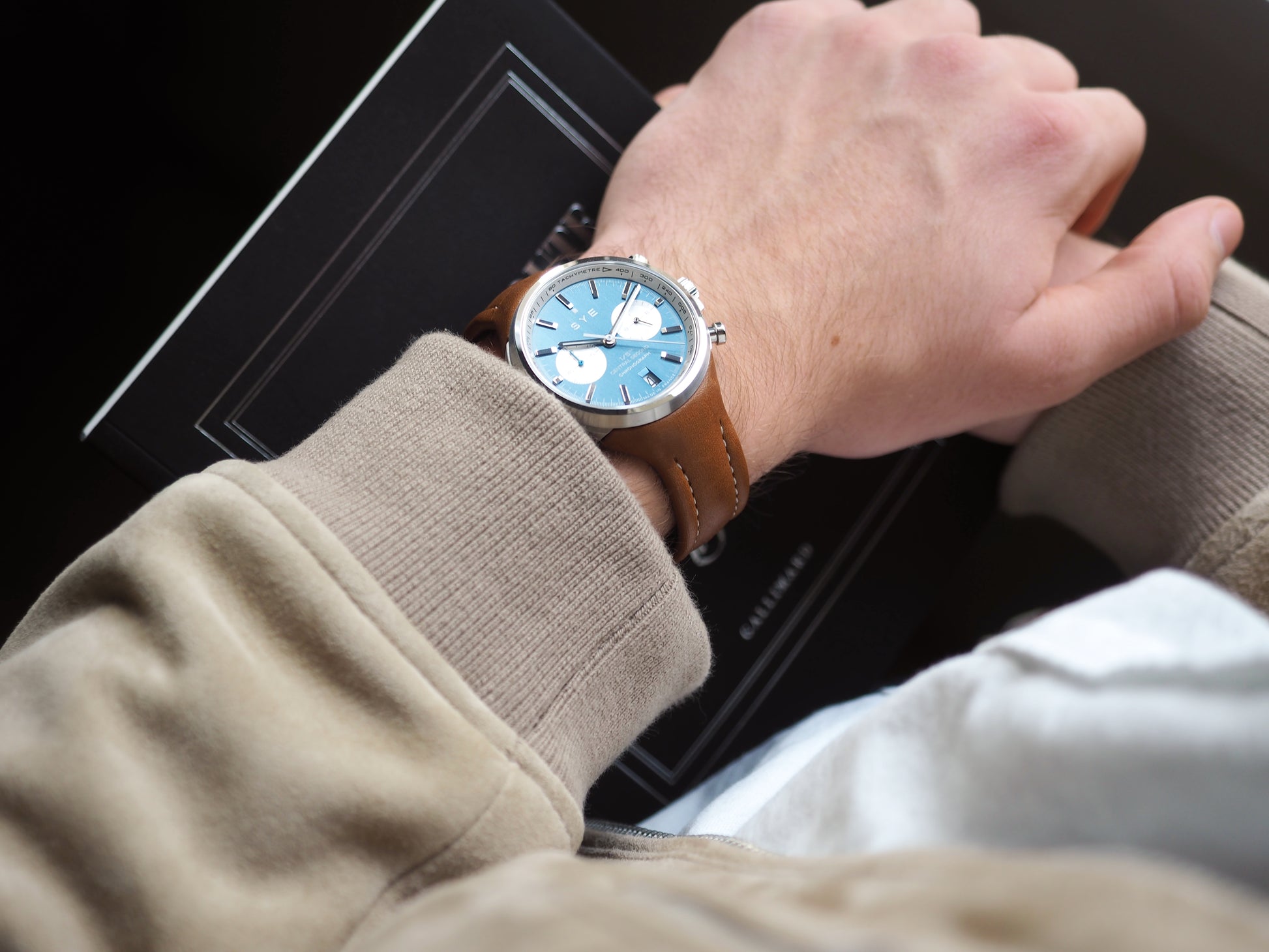 Hand wearing a watch with a blue face and brown leather strap, sitting on a car dashboard.