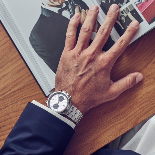 Hand wearing a silver watch on a wooden surface with a magazine in the background