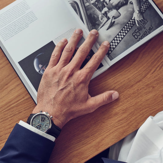 Hand wearing a watch on a wooden surface with an open book featuring black and white photos.