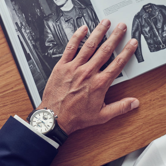 Hand wearing a watch on a wooden surface with a magazine open to a leather jacket page.