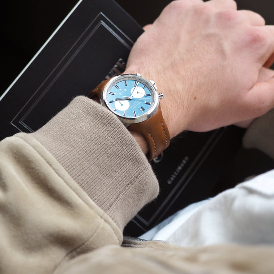 Hand wearing a watch with a blue face and brown leather strap, sitting on a car dashboard.