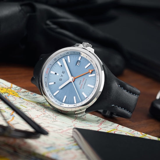 Watch with a blue face and black strap on a map with a pen and glasses.
