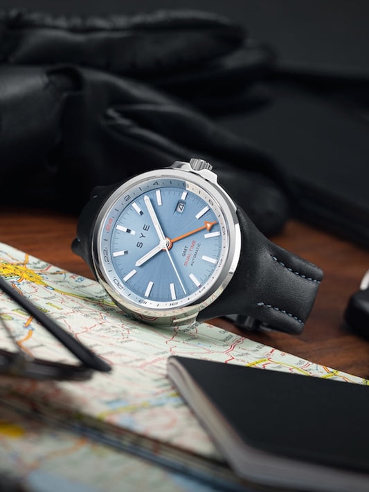 Watch with a blue face and black strap on a map with a pen and glasses.