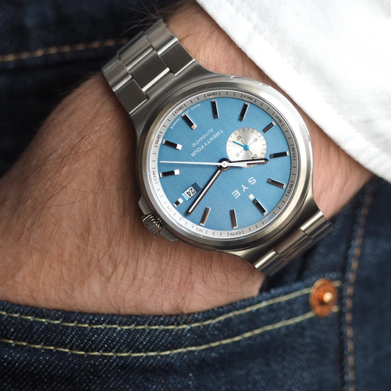 Silver watch with a blue face on a wrist, wearing denim jeans.