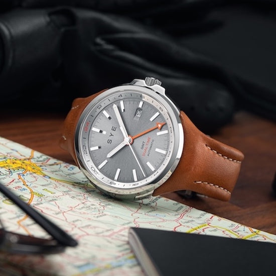 Watch with brown leather strap on a map with gloves and pen in the background